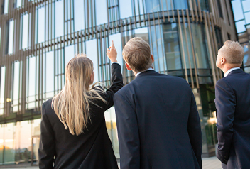 Three people in business suits stand outside, looking up at the glass facade of a modern office building; one person points upward toward the structure.