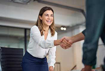 Smiling woman in business attire leans forward to shake hands with another person in an office setting, suggesting a greeting or agreement.