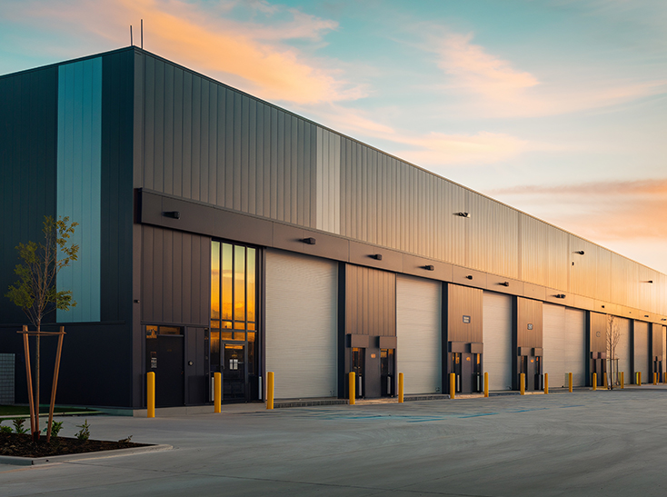 A modern, industrial warehouse with multiple large loading bay doors and yellow safety bollards, set under a colorful sky at sunrise or sunset. The paved lot in front is empty.