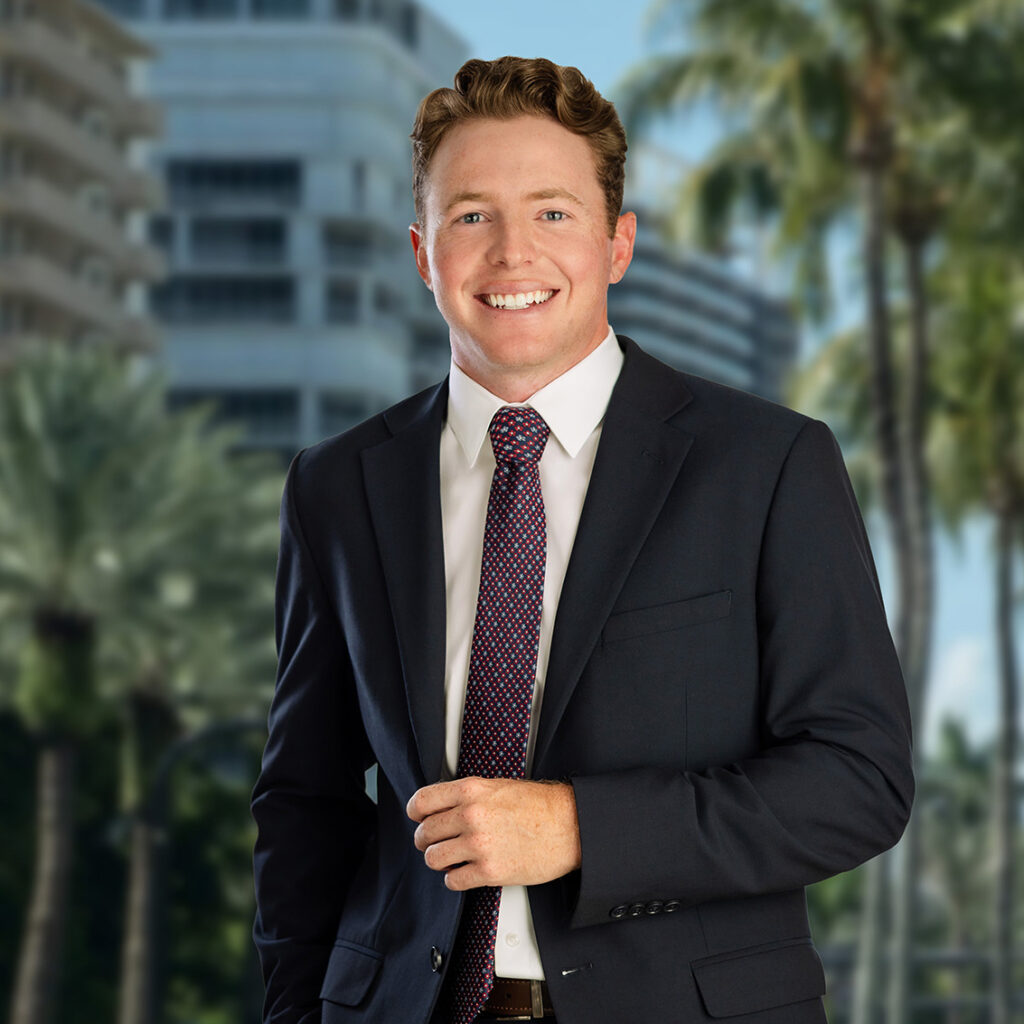 A young man in a dark suit and patterned tie smiles confidently, standing outdoors with modern buildings and palm trees blurred in the background.