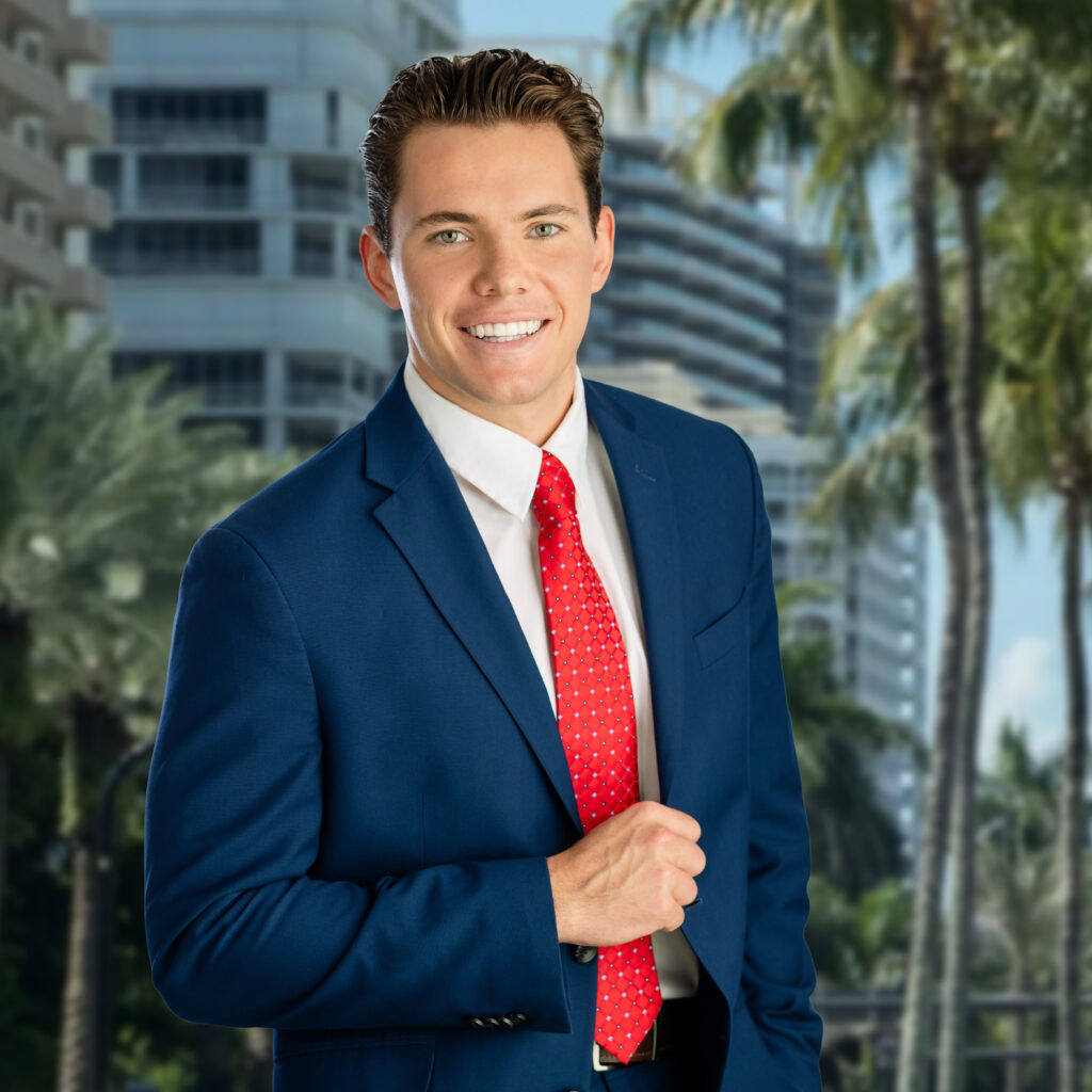 A young man in a blue suit, white shirt, and red tie smiles confidently while standing outdoors in front of palm trees and modern buildings on a sunny day.