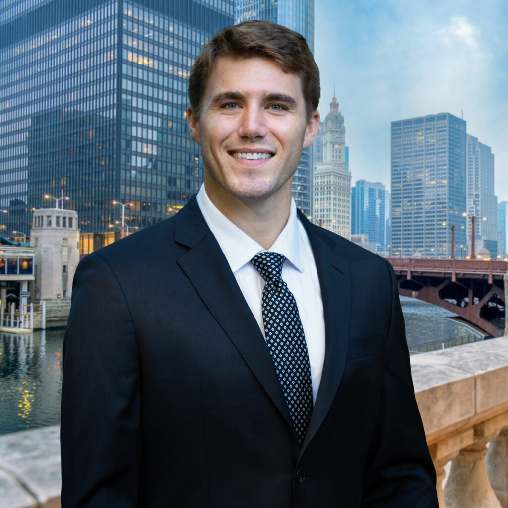 A man in a dark suit and tie smiles while standing outdoors with tall modern buildings, a river, and a bridge in the background, likely in an urban city setting.
